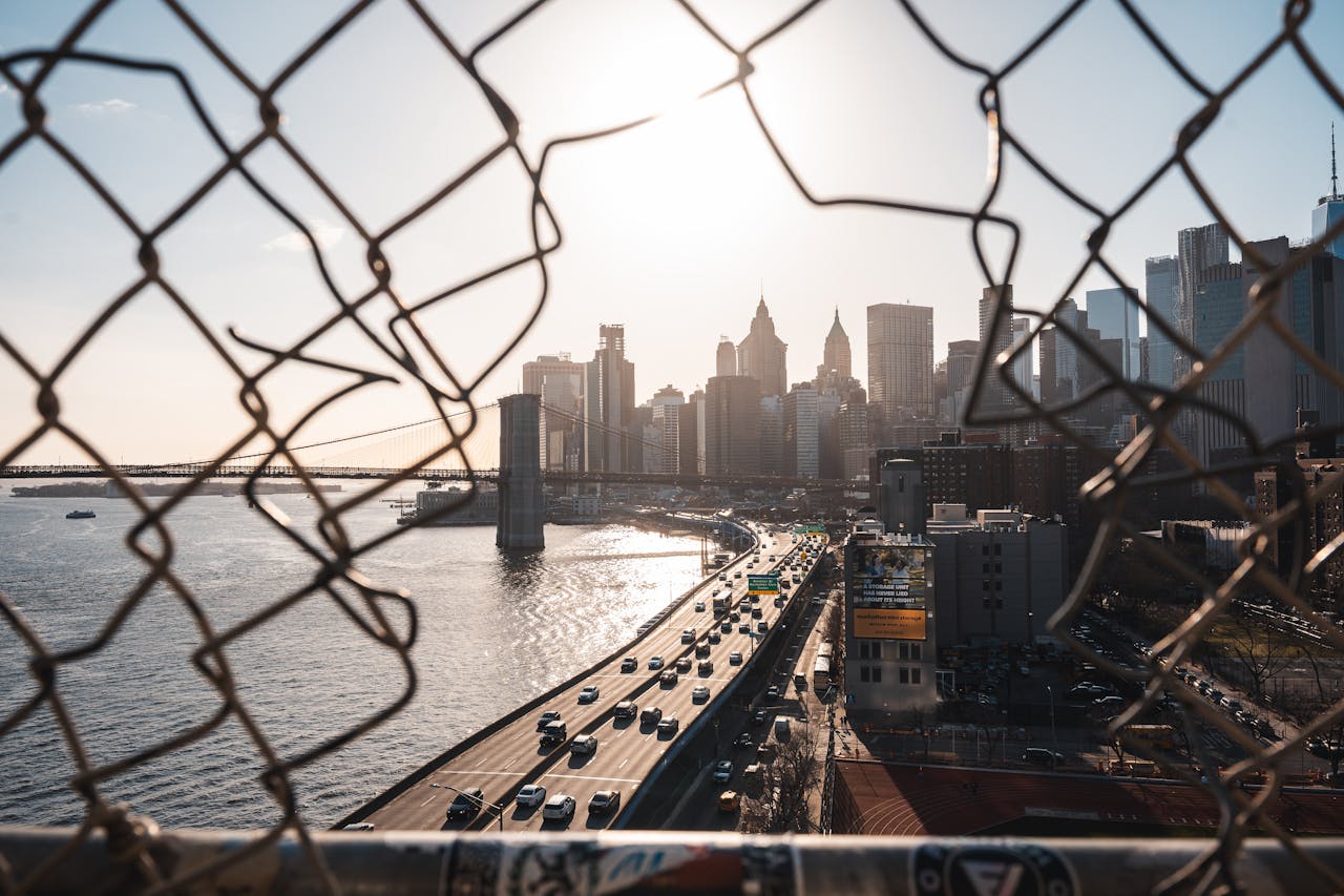 services-04 A view of the New York City skyline through a fence with a bridge and roadway below, capturing urban life.